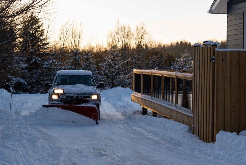 Winter scene of a pickup’s blade scraping snow from a drive, with a new deck and fence at the side