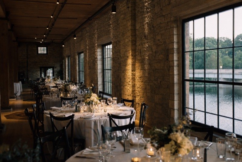 Dinner tables in white linen down a limestone-walled hall, tall windows looking over a river
